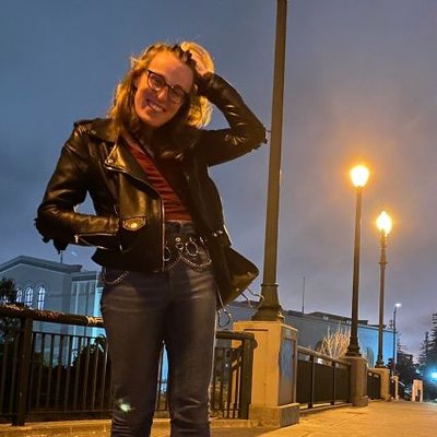joan - a white, queer and trans woman - stands, holding back her dirty-blonde hair and smiling at the camera.